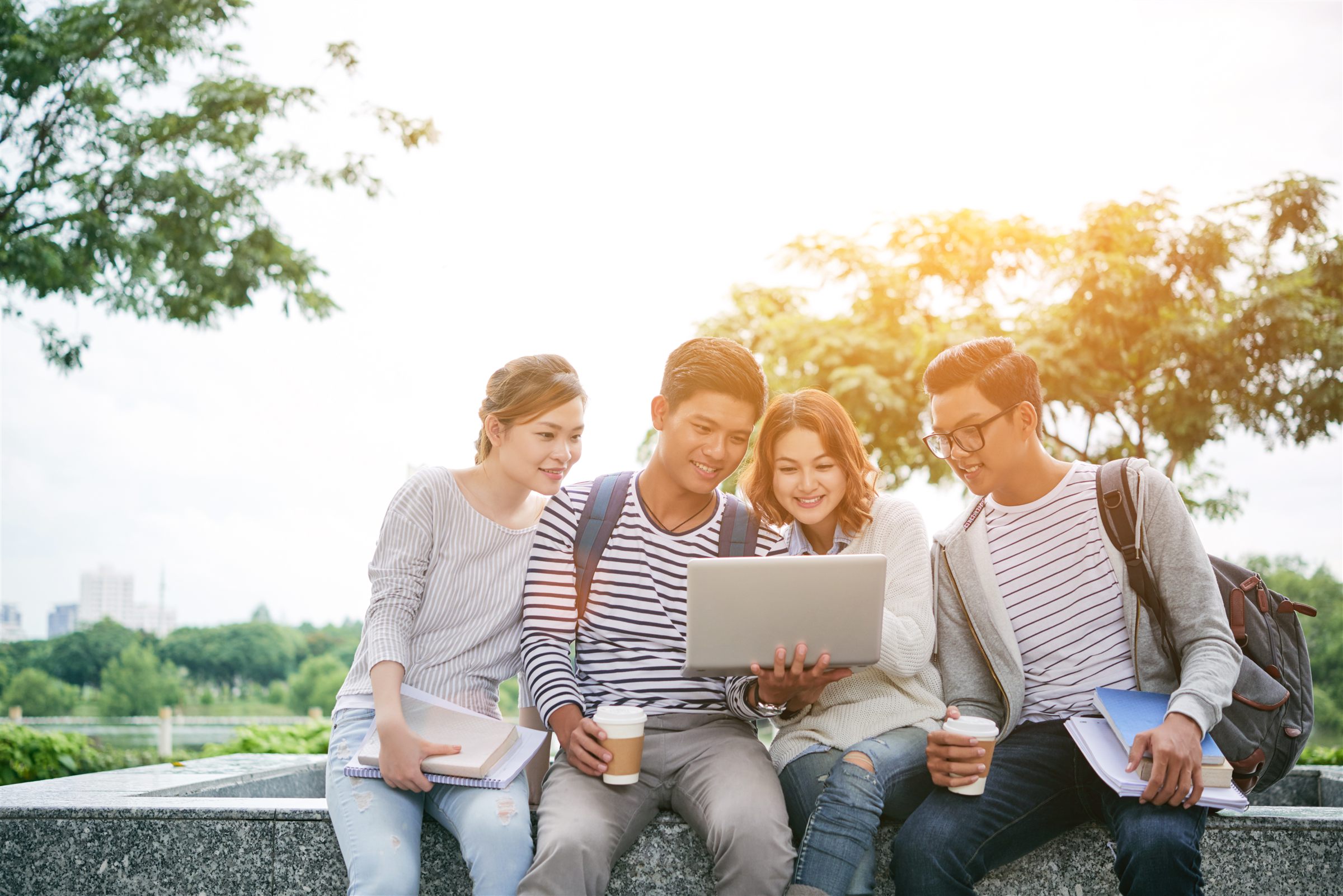 Four young Vietnamese students sitting outdoors on a stone ledge, smiling at a laptop together, with coffee cups and notebooks