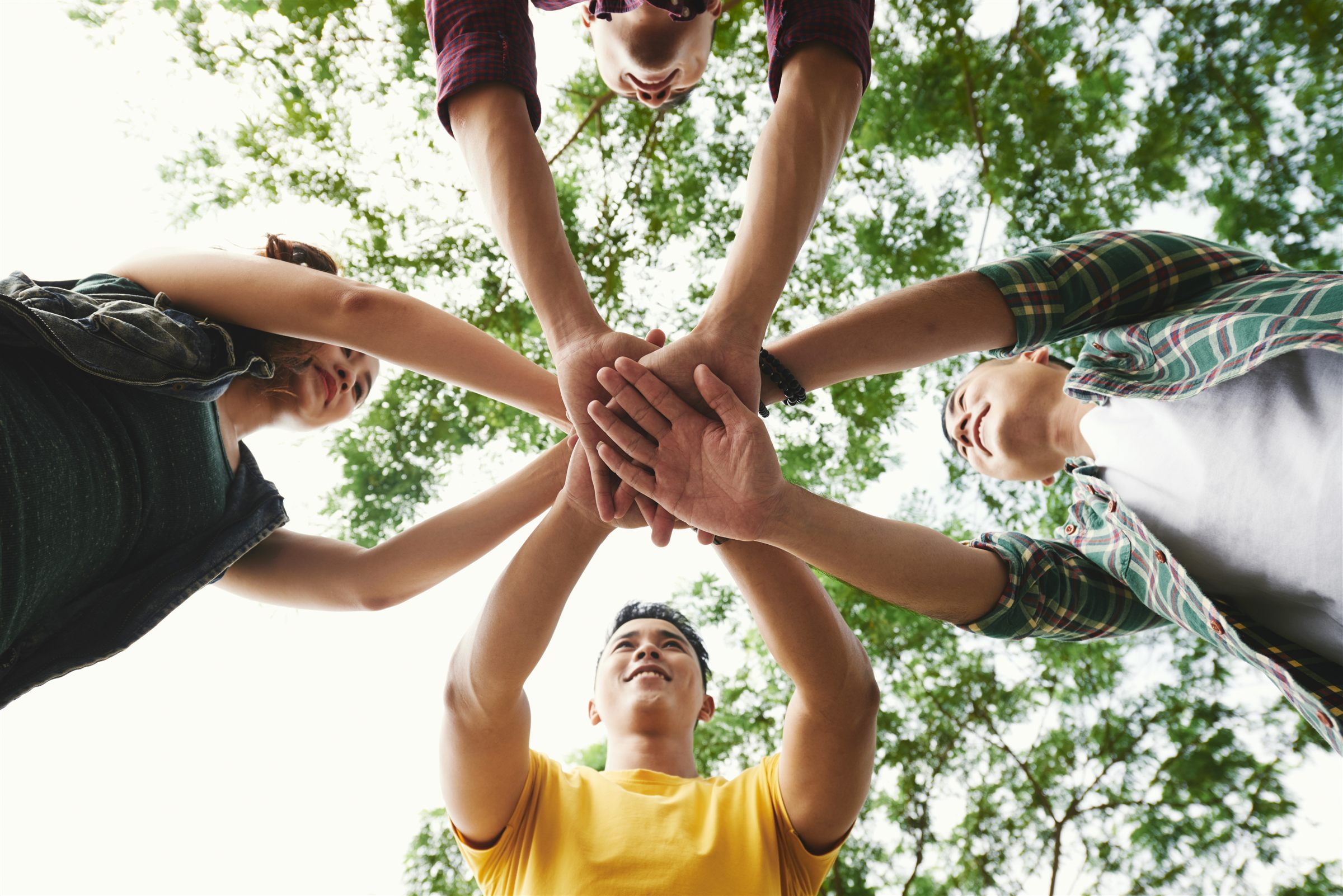 A circle of young people stacking their hands together, seen from below against the trees
