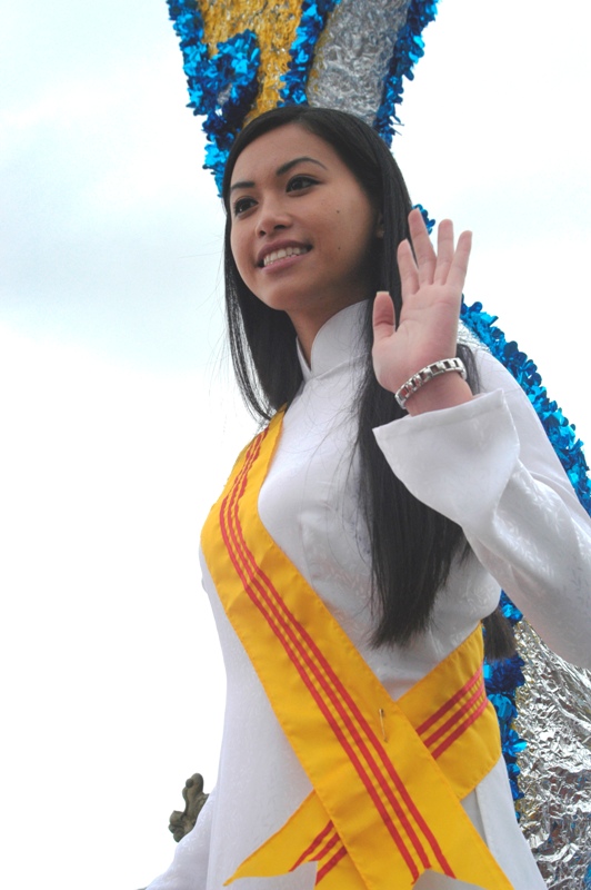 A young woman in a white áo dài with a yellow and red sash waves from a parade float